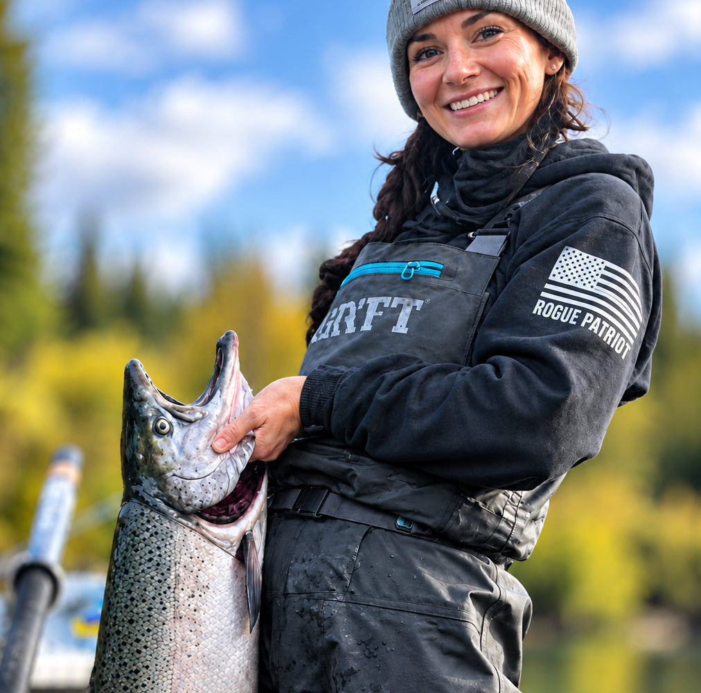 A woman in a boat on the water wearing a gray beanie, a black hoodie with rogue patriot American flag on the sleeve , and black waders, holding a large salmon fish.