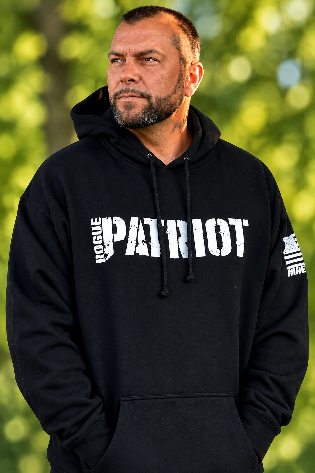A man wearing a black hoodie with 'ROGUE PATRIOT' printed in white across the chest, standing outdoors in front of a tree.