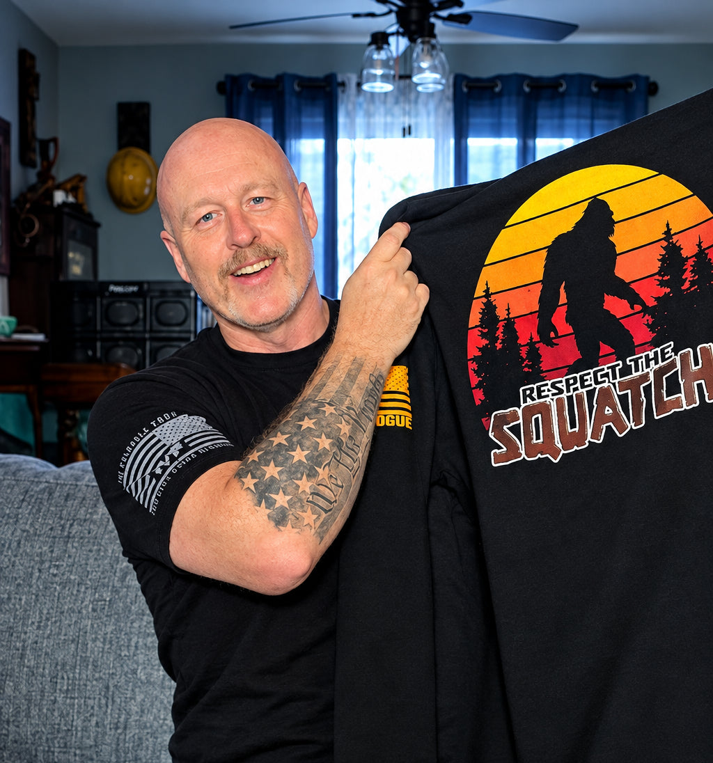 A man holding up a black t-shirt in a living room setting. The shirt features a large graphic print of a Bigfoot silhouette against a sunset gradient with the text 'RESPECT THE SQUATCHI'.