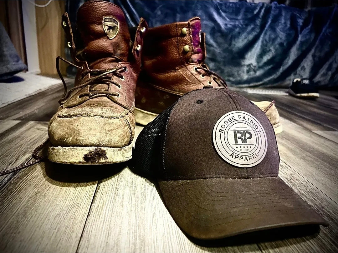 A pair of worn brown leather lace-up work boots and a dark brown trucker hat with a circular 'Rogue Apparel' patch, placed on a wooden floor with a blue fabric backdrop.