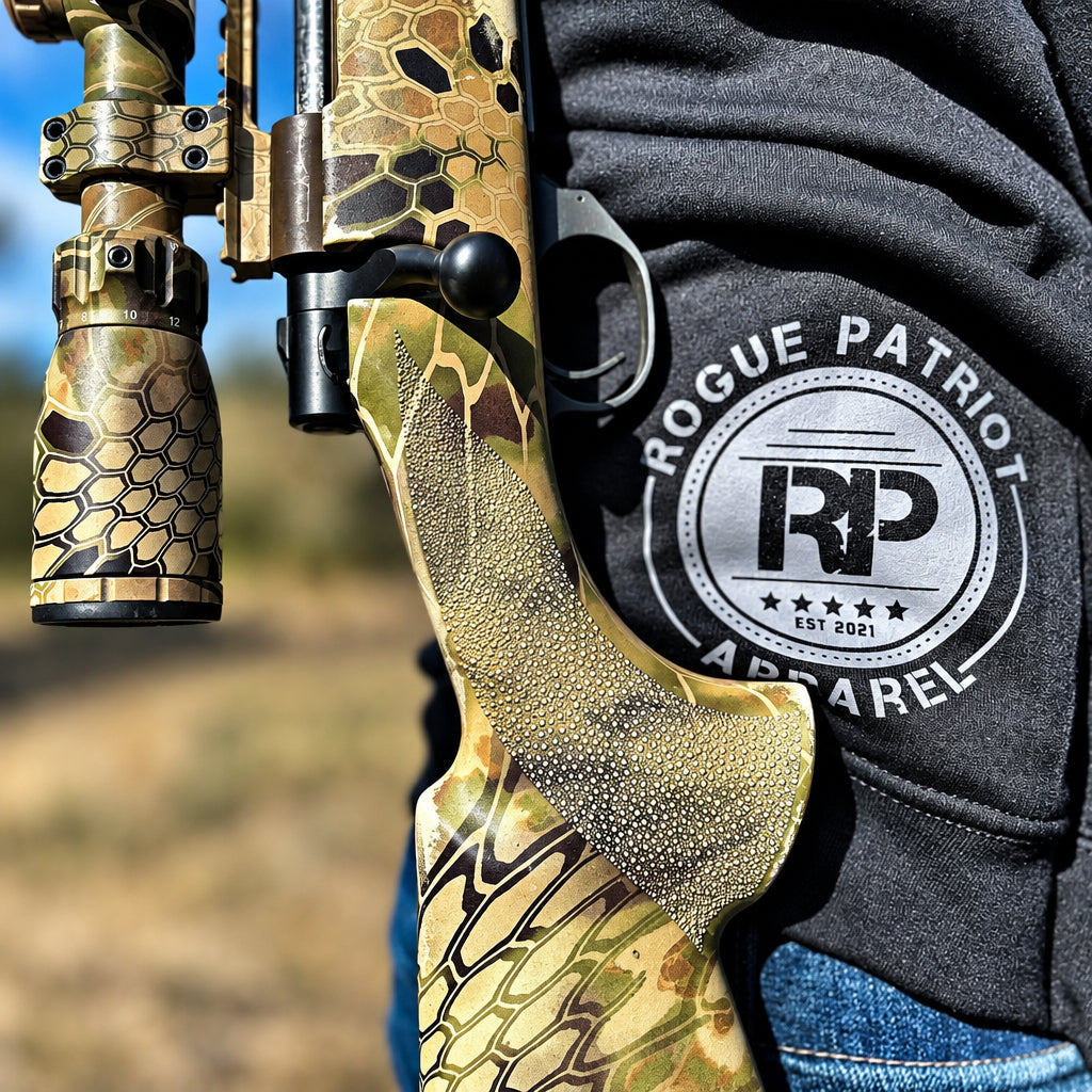 Close-up of a rifle with a tan and black honeycomb-patterned stock and scope, worn on a person's shoulder next to a black hoodie featuring a circular 'Rogue Patriot Apparel' logo.