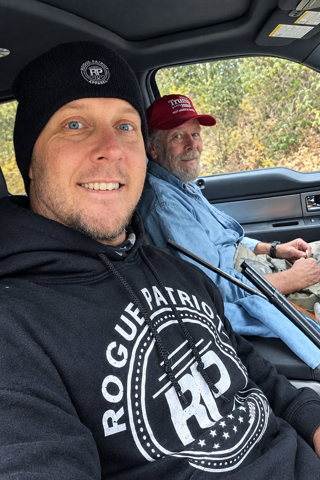 A man in a black beanie and black hoodie with 'ROGUE PATRIOT APPAREL' printed on it sits in the front passenger seat of a vehicle, smiling at the camera. Another man wearing a red cap and light blue shirt sits behind 