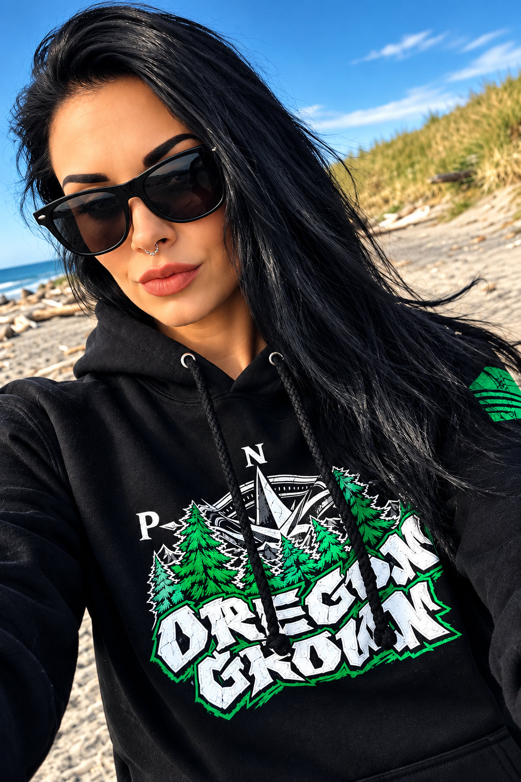 A woman wearing a black hoodie with a green and white 'Oregon Grown' graphic, photographed in a selfie-style close-up on a beach.