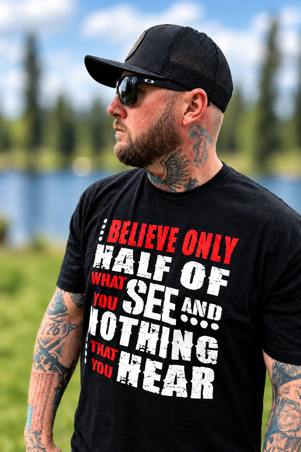 A man wearing a black graphic t-shirt with a humorous quote, a black baseball cap, and sunglasses, standing outdoors near a body of water.