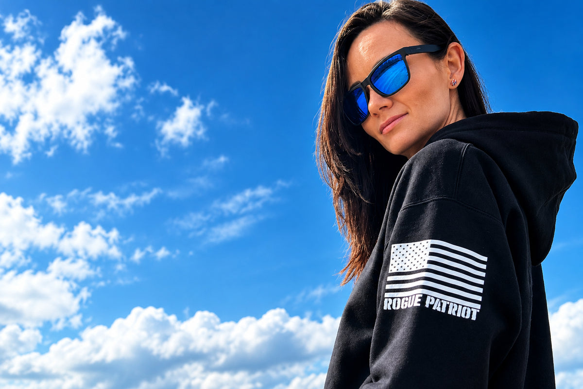 A woman wearing a black hoodie with a white distressed American flag graphic and the text 'ROGUE PATRIOT' on the sleeve, photographed outdoors against a blue sky.
